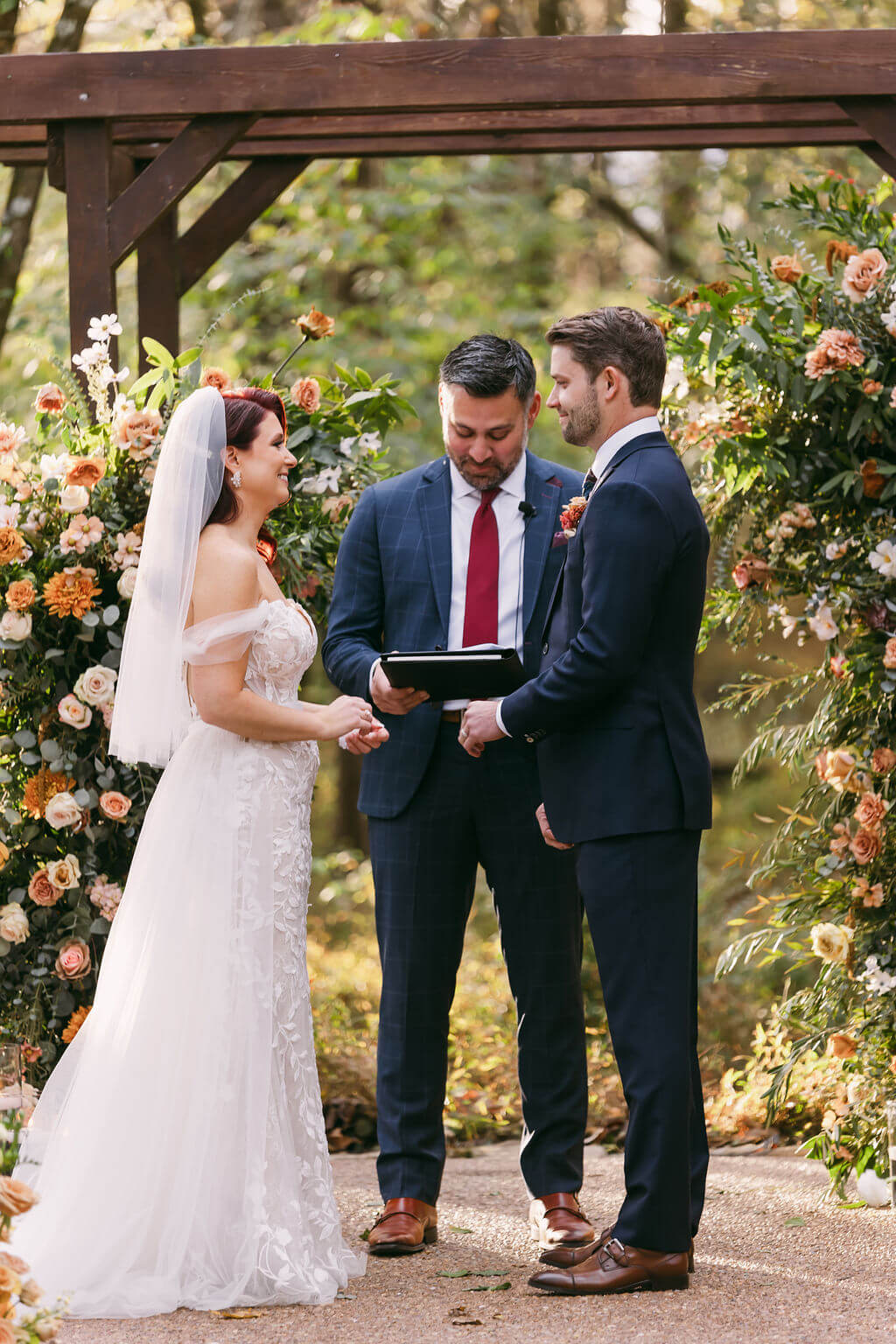 The bride and groom are outside in front of a rustic wooden arch covered in flowers
